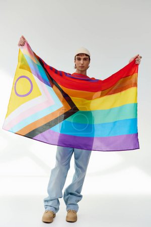 Téléchargez les photos : Joyeux attrayant gay homme en tenue vive avec chapeau blanc tenant drapeau arc-en-ciel et regardant caméra - en image libre de droit