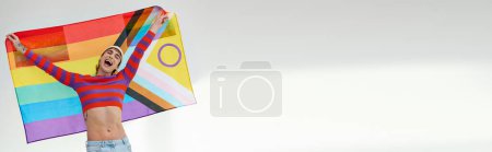 Photo for Merry gay man in vivid outfit with white hat holding rainbow flag and looking at camera, banner - Royalty Free Image