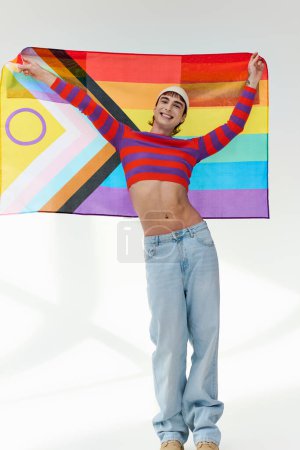 Téléchargez les photos : Positif attrayant gay homme en tenue vive avec chapeau blanc tenant drapeau arc-en-ciel et regardant caméra - en image libre de droit