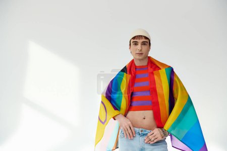 Téléchargez les photos : Joyeux bel homme gay en tenue vive avec chapeau blanc tenant drapeau arc-en-ciel et regardant la caméra - en image libre de droit