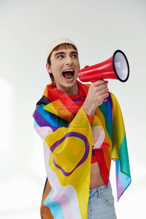 Téléchargez les photos : Joyeux élégant gay homme en tenue vibrante avec drapeau arc-en-ciel en utilisant mégaphone et détourner les yeux - en image libre de droit