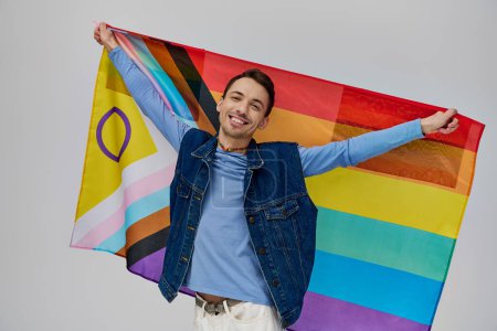 Téléchargez les photos : Joyeux attrayant gay homme dans vibrant casual tenue tenant drapeau arc-en-ciel et souriant à la caméra - en image libre de droit