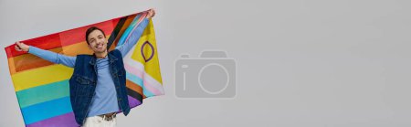 Téléchargez les photos : Joyeux attrayant gay homme dans vibrant casual tenue tenant drapeau arc-en-ciel et souriant à la caméra, bannière - en image libre de droit