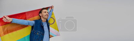 Foto de Alegre guapo gay hombre en moderno traje sosteniendo arco iris bandera y mirando lejos, orgullo mes, bandera - Imagen libre de derechos