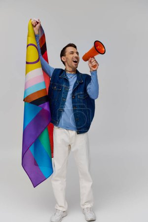 Foto de Joven bien parecido gay hombre en acogedor atuendo sosteniendo arco iris bandera y el uso de megáfono en gris telón de fondo - Imagen libre de derechos