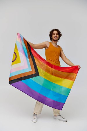 Foto de Positivo seductor gay hombre con oscuro pelo sosteniendo arco iris bandera y sonriendo felizmente a cámara - Imagen libre de derechos