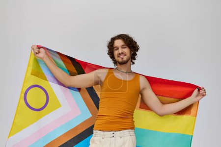Foto de Contento seductor gay hombre con oscuro pelo celebración arco iris bandera y sonriendo felizmente a cámara - Imagen libre de derechos