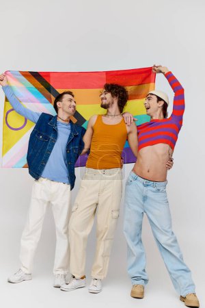 Téléchargez les photos : Trois séduisants gays gays dans des vêtements confortables posant activement avec drapeau arc-en-ciel sur fond gris - en image libre de droit