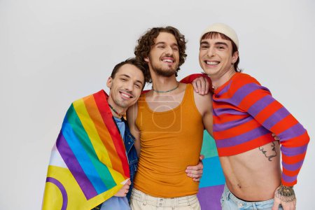 Photo for Three joyous stylish gay men in cozy clothing posing actively with rainbow flag on gray backdrop - Royalty Free Image