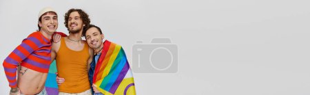 Foto de Tres alegre gay los hombres en acogedor ropa posando activamente con arco iris bandera en gris telón de fondo, bandera - Imagen libre de derechos