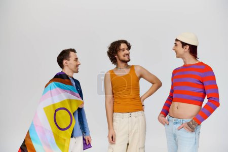 Foto de Tres guapo alegre gay los hombres en acogedor ropa posando activamente con arco iris bandera en gris telón de fondo - Imagen libre de derechos