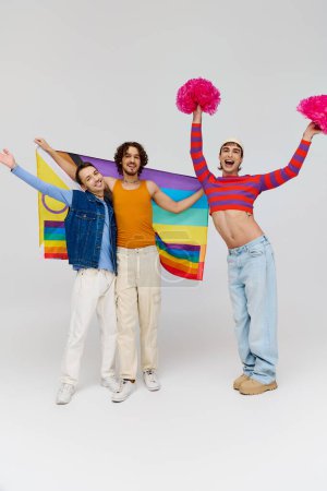 positivo atractivo gay los hombres en vibrante ropa posando con arco iris bandera y pompones en gris telón de fondo