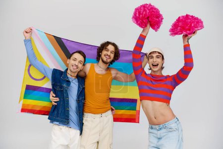 fröhlich ansprechende schwule Männer in lebendigen Kleidern posieren mit Regenbogenfahne und Bommelmützen vor grauem Hintergrund