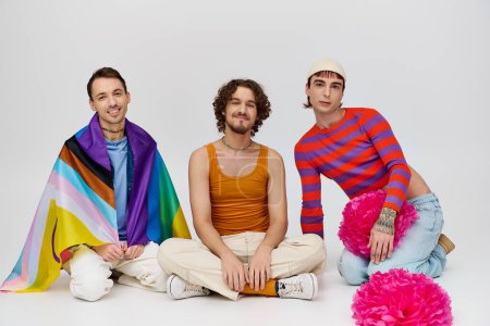 alegre atractivo gay los hombres en vibrante ropa posando con arco iris bandera y pompones en gris telón de fondo