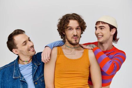 positive junge schwule Männer in lebendigen Kleidern posieren zusammen vor grauem Hintergrund und schauen in die Kamera