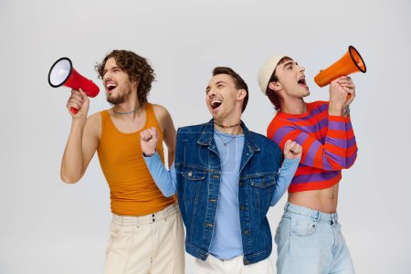 three handsome jolly gay men in vibrant attires using megaphones while posing on gray background