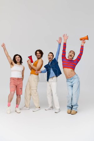 four cheerful trendy gay men in stylish outfits using megaphones and looking at camera happily