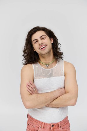 Téléchargez les photos : Joyeux attrayant gay homme avec de longs cheveux en tenue décontractée souriant à la caméra sur fond gris - en image libre de droit