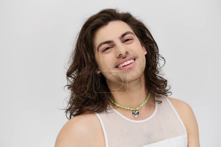 Téléchargez les photos : Joyeux attrayant gay homme avec de longs cheveux en tenue décontractée souriant à la caméra sur fond gris - en image libre de droit