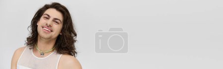 Photo for Merry appealing gay man with long hair in casual attire smiling at camera on gray backdrop, banner - Royalty Free Image