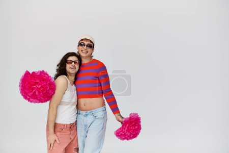 atractivo alegre gay los hombres en vibrantes atuendos con gafas de sol posando con pompones en gris telón de fondo