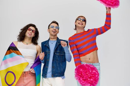drei fröhliche, stylische schwule Männer in fetter Kleidung mit Sonnenbrille posieren mit Bommelmützen und Regenbogenfahne