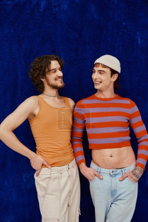Photo for Two joyous good looking gay men in vibrant clothes posing on dark blue backdrop, pride month - Royalty Free Image
