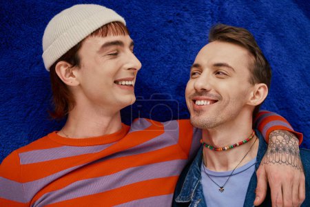 Foto de Dos alegre bien parecido gay los hombres en vibrante ropa posando en oscuro azul telón de fondo, orgullo mes - Imagen libre de derechos