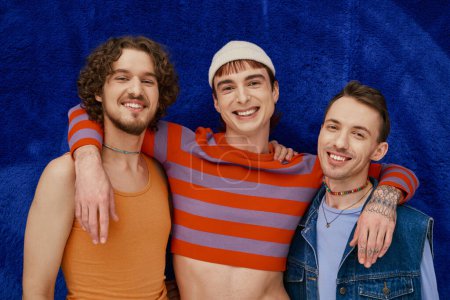 three modish appealing merry gay friends in cozy attires posing on dark blue backdrop, pride month