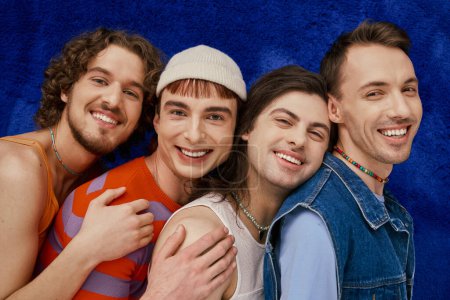 vier fröhlich ansprechende stilvolle schwule Freunde in alltäglichen hellen Kleidern vor blauem Hintergrund, stolzer Monat