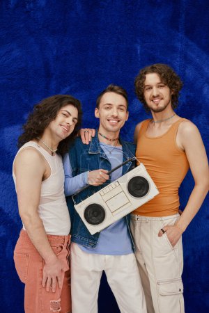 three modish joyous gay friends posing with tape recorder on dark blue backdrop, pride month