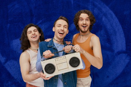 three young joyous gay friends posing with tape recorder on dark blue backdrop, pride month