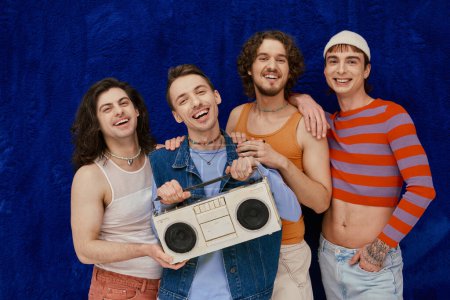 four voguish joyous gay friends posing with tape recorder on dark blue backdrop, pride month