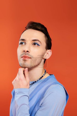 Foto de Alegre atractivo elegante gay hombre con vibrante maquillaje posando en naranja telón de fondo, orgullo mes - Imagen libre de derechos