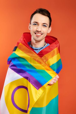 positiv ansprechende junge schwule Männer in kuscheliger Kleidung mit Regenbogenfahne auf orangefarbenem Hintergrund, stolzer Monat