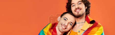 Téléchargez les photos : Joyeux attrayants hommes gays en vêtements confortables avec drapeau arc-en-ciel sur fond orange, mois de fierté, bannière - en image libre de droit