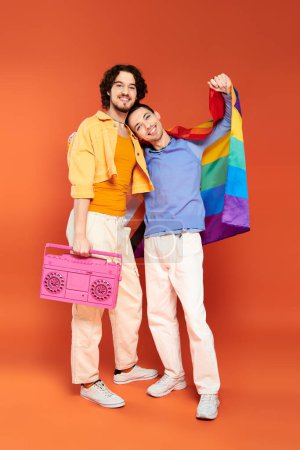 Photo for Two positive handsome gay friends posing with tape recorder and rainbow flag on orange backdrop - Royalty Free Image