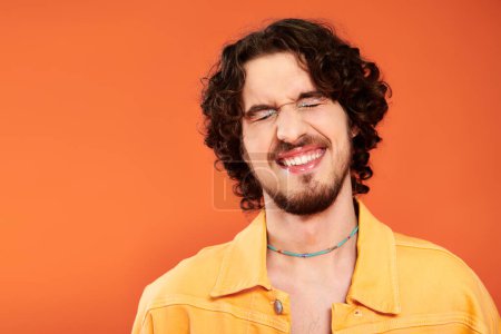 Foto de Feliz atractivo gay hombre con oscuro cabello y vibrante maquillaje posando en naranja telón de fondo, orgullo mes - Imagen libre de derechos