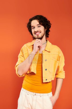 Foto de Contento atractivo gay hombre con oscuro cabello y vibrante maquillaje posando en naranja telón de fondo, orgullo mes - Imagen libre de derechos