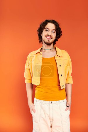 positiv ansprechender homosexueller Mann mit dunklen Haaren und lebendigem Make-up, der auf orangefarbenem Hintergrund posiert, Monat des Stolzes