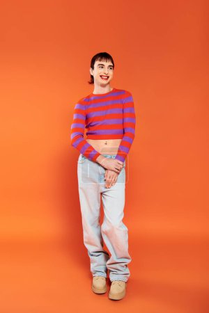 Téléchargez les photos : Joyeux attrayant gay l'homme dans vibrant vêtements avec maquillage posant sur orange toile de fond et regarder loin - en image libre de droit