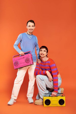 Photo for Two joyful good looking gay men with vibrant makeup posing with tape recorders on orange backdrop - Royalty Free Image