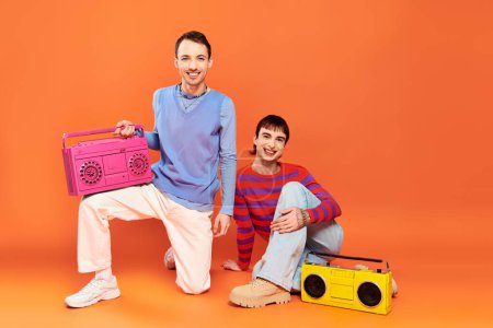 Photo for Two cheerful good looking gay men with vibrant makeup posing with tape recorders on orange backdrop - Royalty Free Image