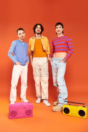 drei positive attraktive schwule Freunde in lebhafter Kleidung posieren mit Tonbandgeräten, stolzer Monat