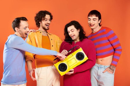 Photo for Four joyous handsome gay friends posing actively with tape recorder on orange backdrop, pride month - Royalty Free Image