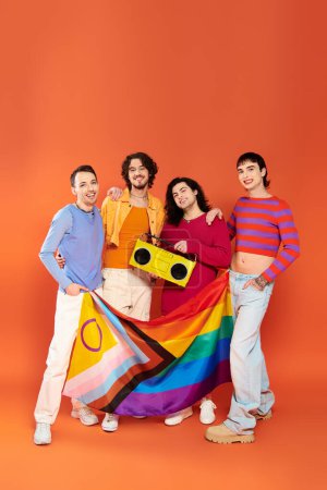Foto de Cuatro jóvenes feliz gay amigos posando con arco iris bandera y cinta grabadora en naranja fondo - Imagen libre de derechos
