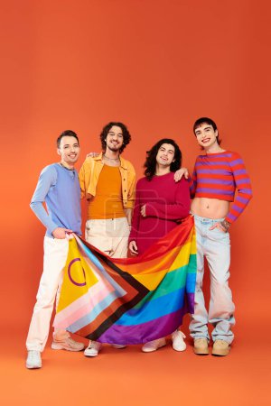 vier positive, modische schwule Männer in lebendigen Kleidern, die Regenbogenfahne vor der Kamera halten, stolz