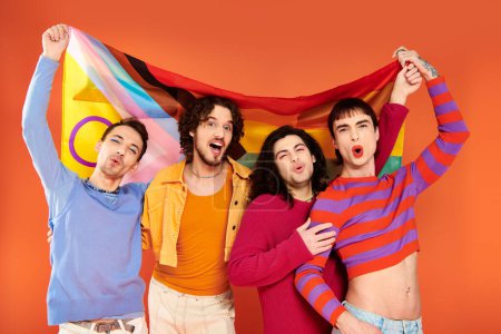 Téléchargez les photos : Quatre gays joyeux à la mode en tenue vibrante tenant le drapeau arc-en-ciel devant la caméra, fierté - en image libre de droit