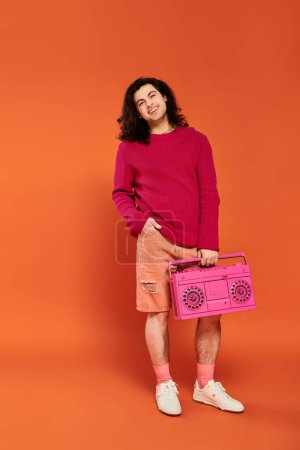 Photo for Jolly appealing gay man in magenta sweatshirt with long hair holding tape recorder smiling at camera - Royalty Free Image
