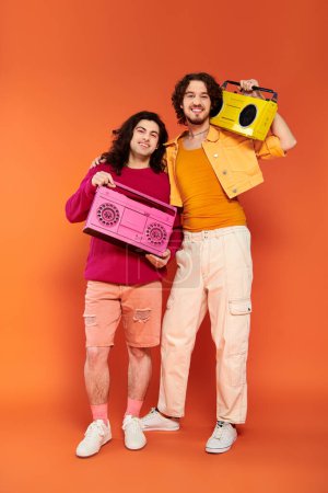 Foto de Dos alegre atractivo gay amigos en vibrante ropa posando con cinta grabadoras, orgullo mes - Imagen libre de derechos